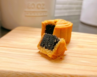 Könnte beinhalten: Zwei Mondkuchen auf einem Holzbrett. Ein Mondkuchen ist halbiert und zeigt eine Füllung aus schwarzem Sesam.