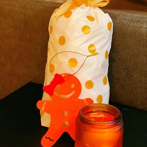 May include: A white gift bag with yellow polka dots and a gold ribbon, a red felt gingerbread person cutout, and a brown glass candle with a warm orange glow.