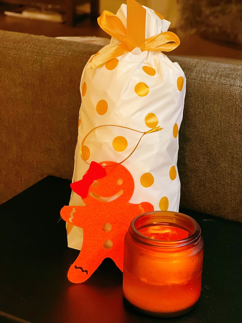 May include: A white gift bag with yellow polka dots and a gold ribbon, a red felt gingerbread person cutout, and a brown glass candle with a warm orange glow.