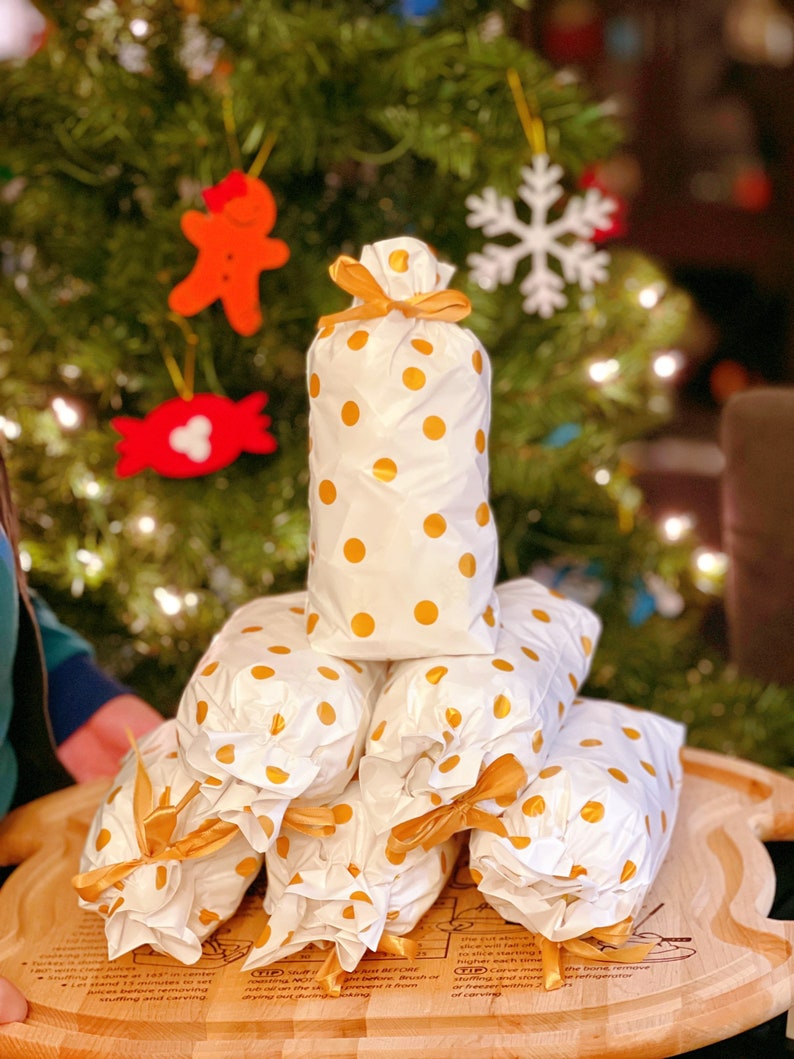 May include: A stack of white gift bags with gold polka dots and gold ribbon bows. The bags are arranged in a pyramid shape on a wooden cutting board. The board has instructions for preparing a turkey.