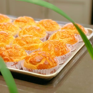 May include: A tray of freshly baked golden brown buns with a crispy, flaky crust. The buns are arranged in rows on a silver tray with white paper liners.