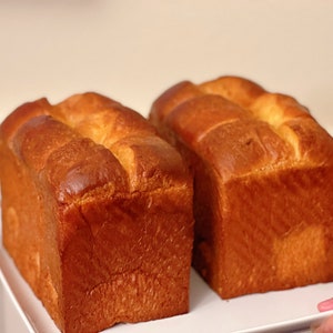 May include: Two golden brown loaves of bread on a white plate. The bread is square shaped and has a slightly crusty exterior.