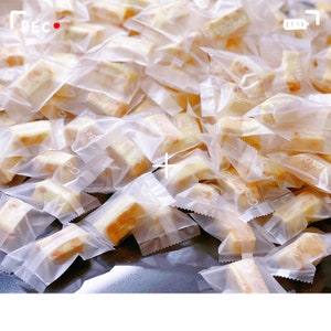 May include: A large pile of individually packaged, translucent, white, rectangular nougat candies. The candies are wrapped in clear plastic bags with white text.