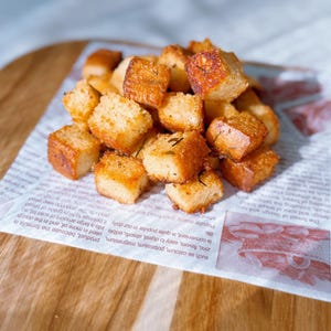 May include: A pile of golden-brown croutons, seasoned with herbs, sits on a piece of newspaper. The croutons are square-shaped and appear crispy. The newspaper is placed on a wooden cutting board.