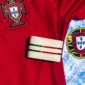 May include: Two thin beaded bracelets featuring a repeating pattern of black, green, and red beads with a single gold-toned bead in the centre, displayed on a cream-coloured textured cuff against a red fabric background with a Portugal crest.