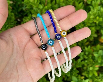 May include: Four beaded bracelets with evil eye charms. The bracelets feature a variety of colors, including gray, turquoise, blue, and yellow. Each bracelet has a white cord and a gold-colored bead. The background is a blurred green.