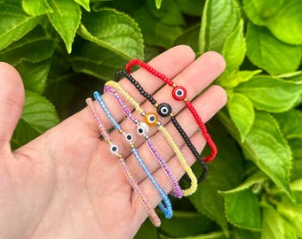 May include: A hand displays six beaded bracelets in various colors: red, black, yellow, purple, blue, and pink. Each bracelet features a small, round evil eye charm with gold accents. The background is a lush green foliage.
