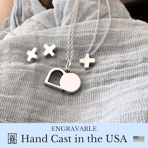 May include: Silver necklace with a heart pendant and a circle charm. The necklace is layered with a smaller chain with a cross charm. Two smaller cross charms are also included. The necklace is engravable and hand cast in the USA.