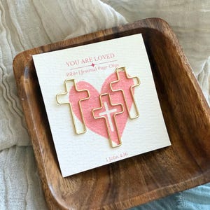 May include: Gold cross-shaped paper clips with the words "You Are Loved" and "Bible | Journal Page Clips" on a white card with a pink heart and the text "1 John 4:16".