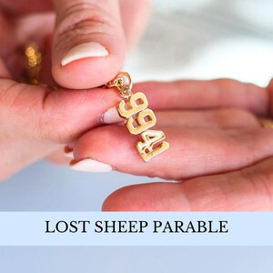 May include: A gold necklace with a pendant that says "1996" in bold, block letters. The necklace is being held by a hand.