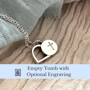 May include: A silver necklace with a heart-shaped pendant and a circular charm featuring a cross. The necklace is displayed on a textured, light-colored fabric. The text "Empty Tomb with Optional Engraving" is visible.
