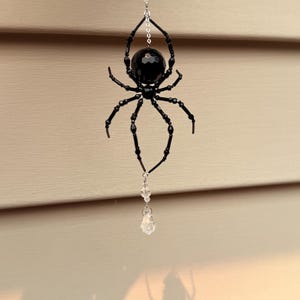 May include: A hanging ornament featuring a black faceted bead spider with eight beaded legs. The spider is suspended from a silver-toned chain accented with clear, faceted crystal beads above and below the body, ending in a clear crystal drop.