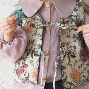 Pu&ograve; includere: Un gilet con stampa floreale con una camicia abbottonata rosa sotto. Il gilet ha una chiusura a cravatta e un bordo blu.