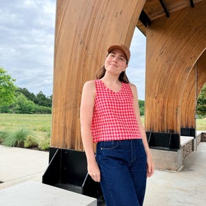 May include: A sleeveless red and pink gingham top paired with dark blue denim jeans. The person is wearing a brown baseball cap. The background features a wooden structure and a cloudy sky.