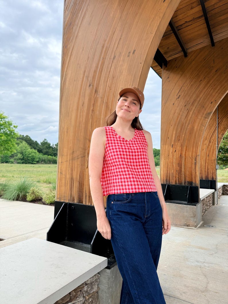 May include: A sleeveless red and pink gingham top paired with dark blue denim jeans. The person is wearing a brown baseball cap. The background features a wooden structure and a cloudy sky.