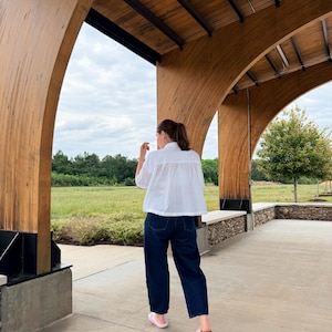 May include: A person wearing a white eyelet shirt, dark blue jeans, and pink slides. The person is standing under a wooden structure with a view of a green field and cloudy sky.