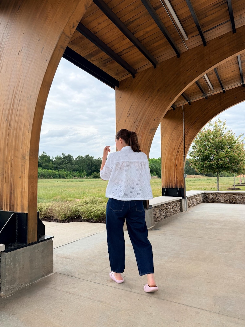 May include: A person wearing a white eyelet shirt, dark blue jeans, and pink slides. The person is standing under a wooden structure with a view of a green field and cloudy sky.
