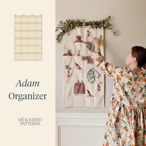 May include: A cream-coloured fabric wall organiser with multiple rows of pockets, featuring a grid diagram and the text Adam Organizer and ME & KIDDO PATTERNS. The pockets contain small doll-sized clothing items and toys, hung with wooden clips.