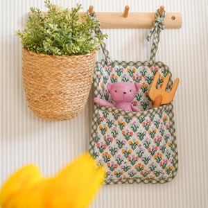 May include: A green and white fabric hanging organizer with a floral print. The organizer has a pocket with a pink teddy bear and a wooden toy rake inside. The organizer is hanging on a wooden hook on a striped wall.