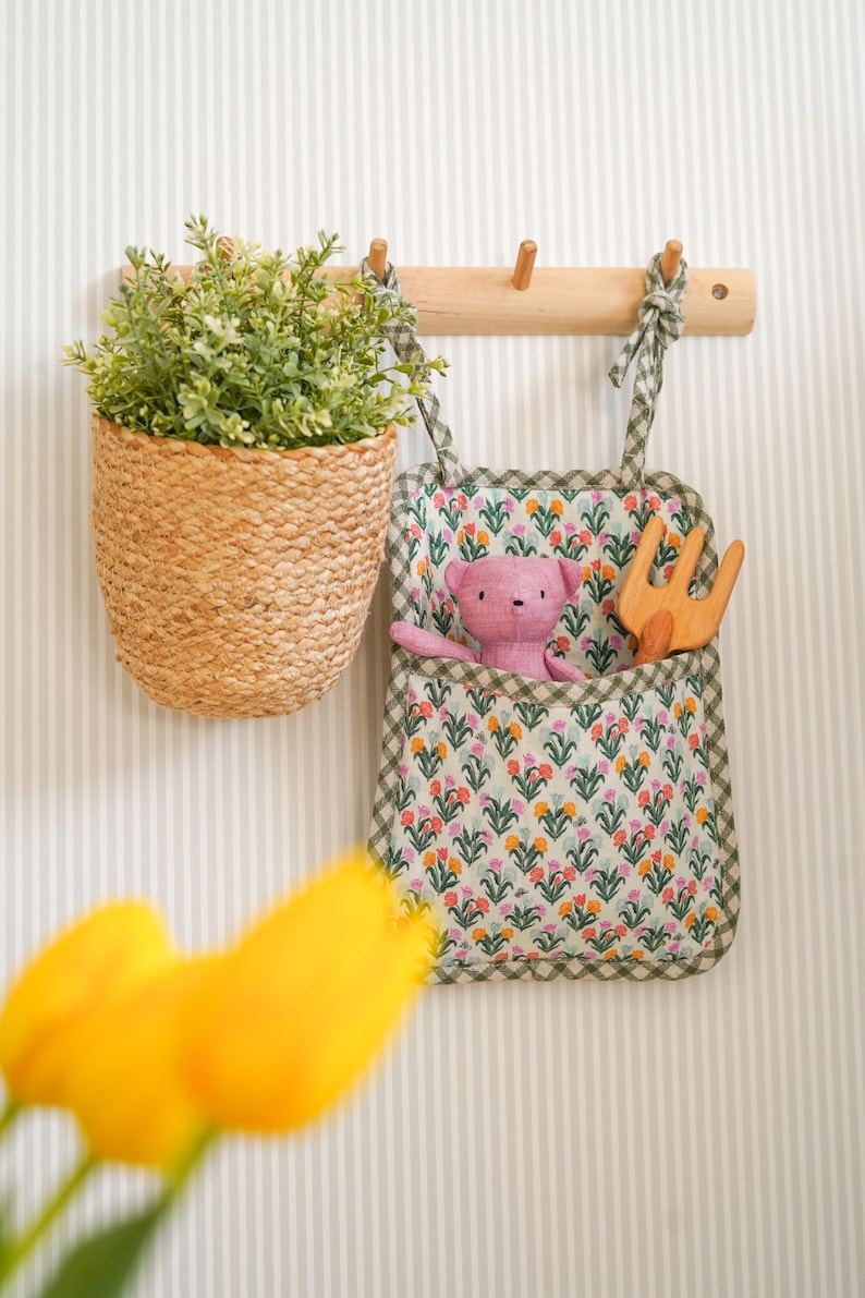 May include: A green and white fabric hanging organizer with a floral print. The organizer has a pocket with a pink teddy bear and a wooden toy rake inside. The organizer is hanging on a wooden hook on a striped wall.