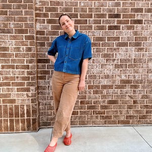 May include: A person wearing a denim blue short-sleeved shirt, tan trousers, and red woven flats. The outfit is casual and the background is a brick wall.
