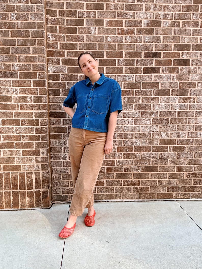 May include: A person wearing a denim blue short-sleeved shirt, tan trousers, and red woven flats. The outfit is casual and the background is a brick wall.
