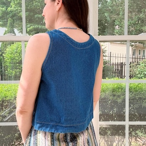 May include: A sleeveless, blue denim top with a rounded neckline. The top is paired with a patterned skirt featuring vertical stripes in various colors. The image is taken indoors, with a window and greenery visible in the background.