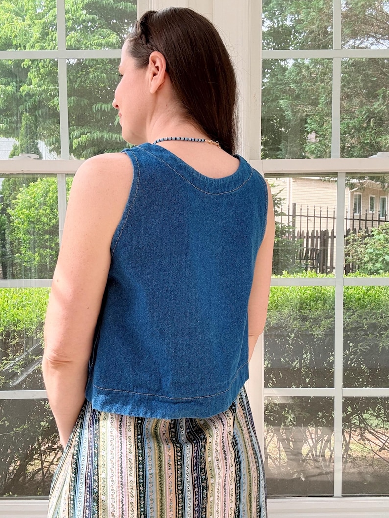 May include: A sleeveless, blue denim top with a rounded neckline. The top is paired with a patterned skirt featuring vertical stripes in various colors. The image is taken indoors, with a window and greenery visible in the background.
