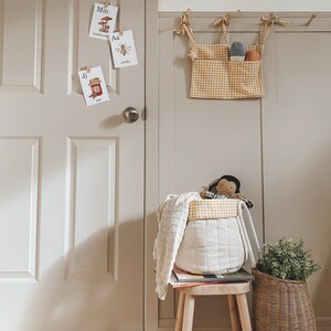 May include: A white and yellow checkered fabric storage bin with a white fabric lining. The bin is hanging on a wall with a white and yellow checkered fabric pocket on the front. The bin is filled with toys, including a doll and a knitted blanket. A small wooden stool with a book on top sits in front of the bin. A wicker basket with a green plant sits on the floor next to the stool.