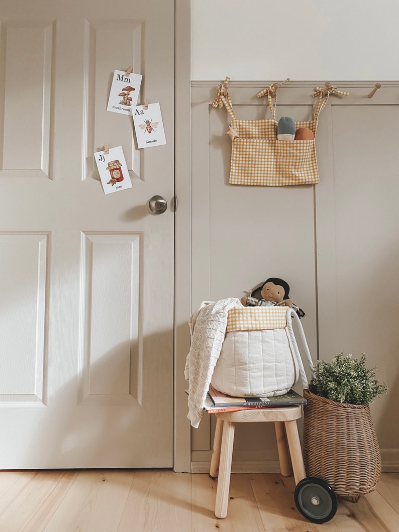 May include: A white and yellow checkered fabric storage bin with a white fabric lining. The bin is hanging on a wall with a white and yellow checkered fabric pocket on the front. The bin is filled with toys, including a doll and a knitted blanket. A small wooden stool with a book on top sits in front of the bin. A wicker basket with a green plant sits on the floor next to the stool.