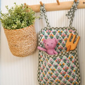 May include: A wooden wall-mounted rack with three hooks, holding a green and white gingham fabric pocket with a pink stuffed animal and a wooden fork, and a woven basket with a green plant.