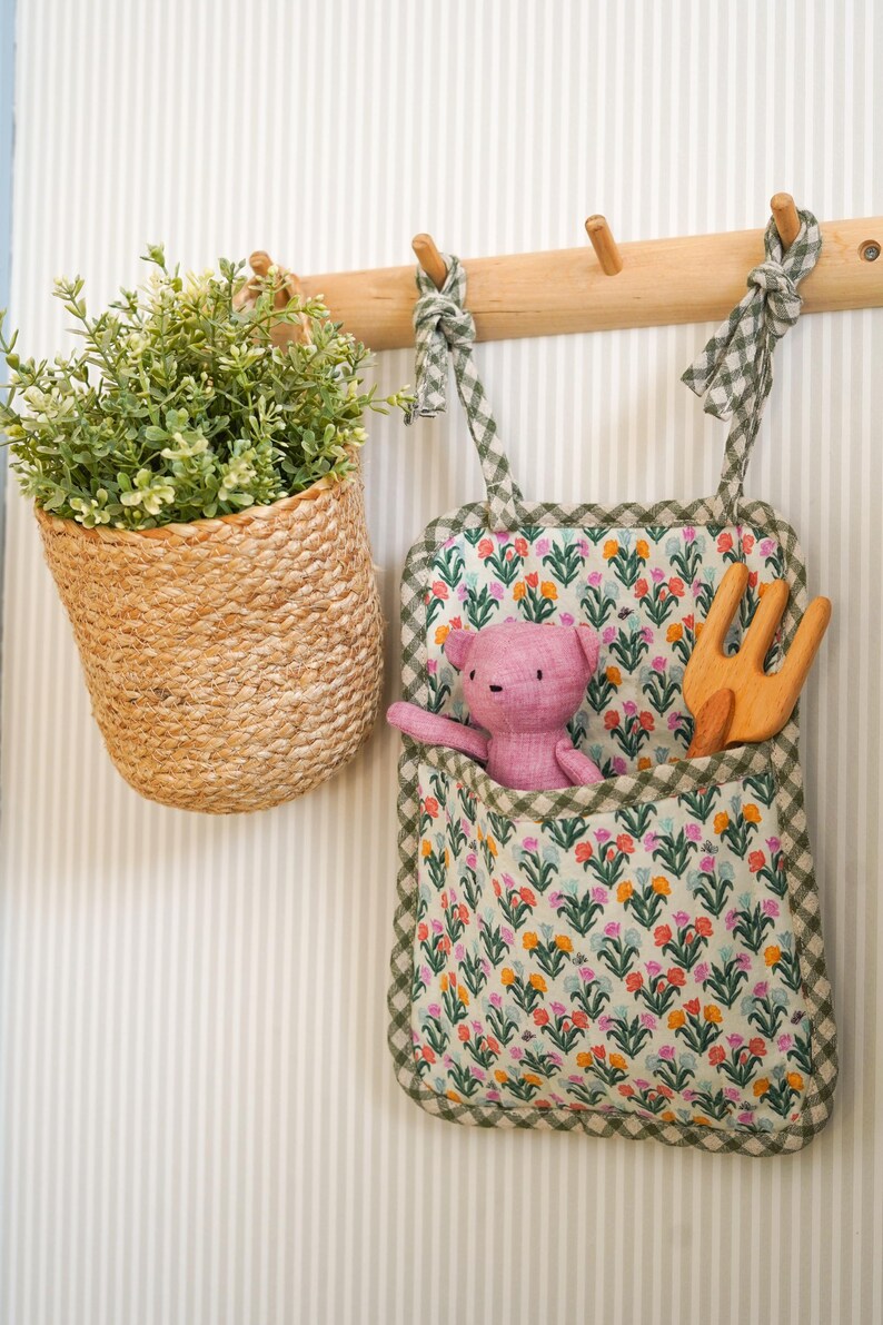 May include: A wooden wall-mounted rack with three hooks, holding a green and white gingham fabric pocket with a pink stuffed animal and a wooden fork, and a woven basket with a green plant.