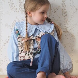 Pu&ograve; includere: Una bambina che indossa una camicia a righe blu e bianche con un grande colletto bianco, un gilet con stampa floreale e pantaloni blu. Ha i capelli in due trecce ed &egrave; seduta su una superficie di legno.
