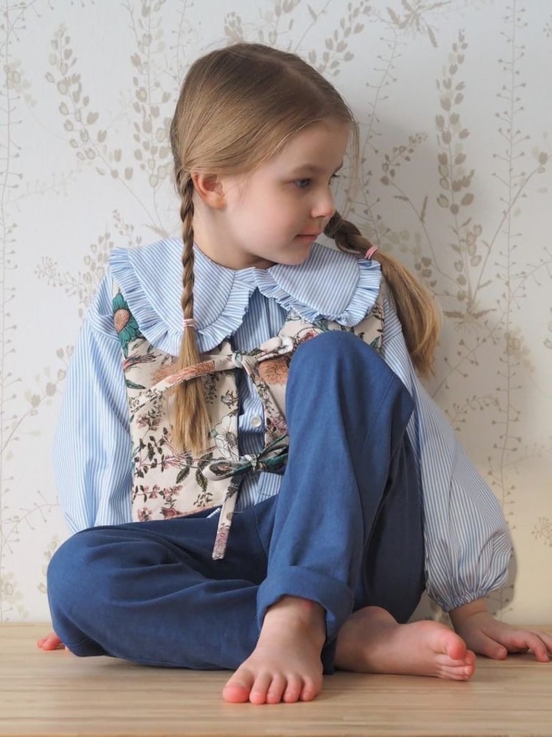 Pu&ograve; includere: Una bambina che indossa una camicia a righe blu e bianche con un grande colletto bianco, un gilet con stampa floreale e pantaloni blu. Ha i capelli in due trecce ed &egrave; seduta su una superficie di legno.