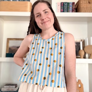 May include: A sleeveless top with a blue and white striped pattern, accented with brown and orange polka dots. The top is paired with beige pants. The overall style is casual and summery.