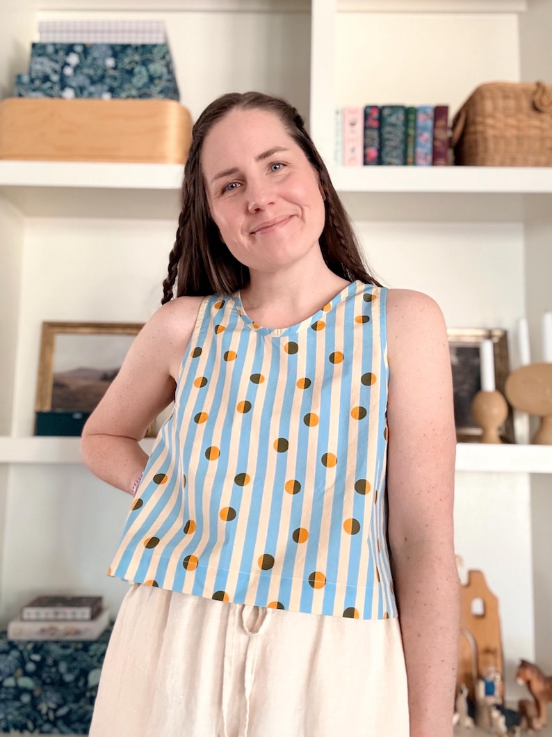 May include: A sleeveless top with a blue and white striped pattern, accented with brown and orange polka dots. The top is paired with beige pants. The overall style is casual and summery.