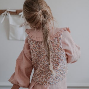 Pu&ograve; includere: Una bambina che indossa una blusa rosa con un gilet a stampa floreale e una gonna marrone. I suoi capelli sono intrecciati.