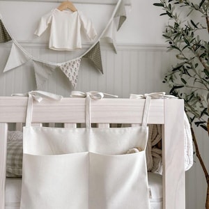 May include: A white canvas crib organizer with two pockets hanging on a white crib. The organizer has white ties to attach it to the crib.