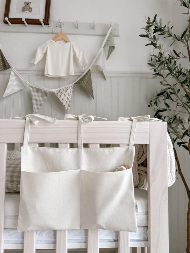 May include: A white canvas crib organizer with two pockets hanging on a white crib. The organizer has white ties to attach it to the crib.