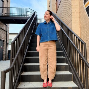 May include: A blue denim short-sleeved shirt with two chest pockets and tan corduroy trousers. The outfit is completed with red woven flats. The person is on a metal staircase with a black railing, outdoors.