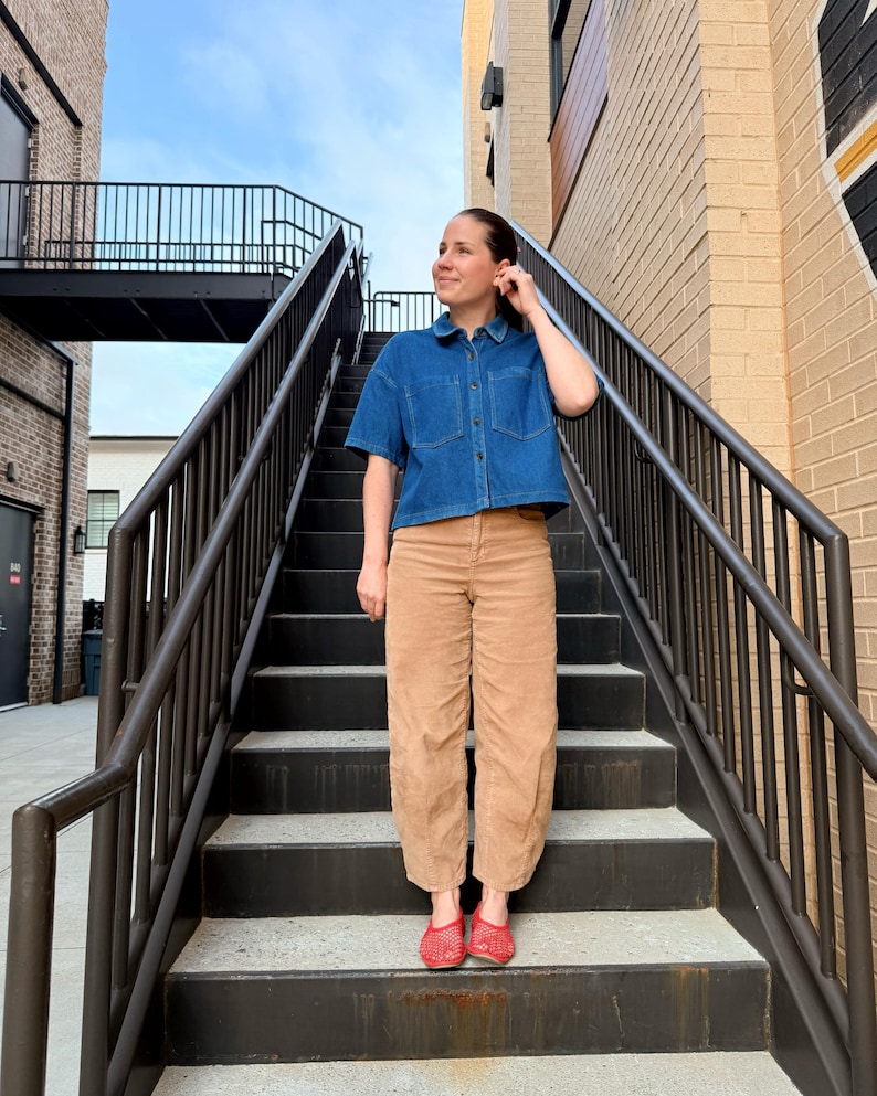 May include: A blue denim short-sleeved shirt with two chest pockets and tan corduroy trousers. The outfit is completed with red woven flats. The person is on a metal staircase with a black railing, outdoors.