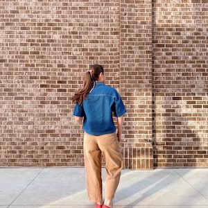 May include: A person wearing a denim blue short-sleeved shirt, tan trousers, and red flats stands in front of a brick wall. The outfit is casual and comfortable, suitable for everyday wear. The brick wall provides a textured backdrop.