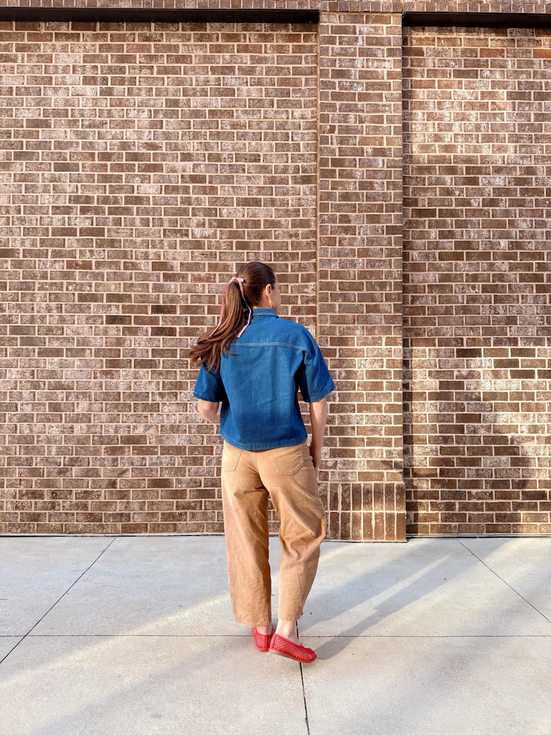 May include: A person wearing a denim blue short-sleeved shirt, tan trousers, and red flats stands in front of a brick wall. The outfit is casual and comfortable, suitable for everyday wear. The brick wall provides a textured backdrop.