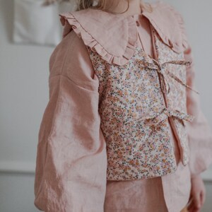 Pu&ograve; includere: Una blusa rosa con un grande colletto a balze e un gilet a stampa floreale. Il gilet ha un nodo sul davanti.