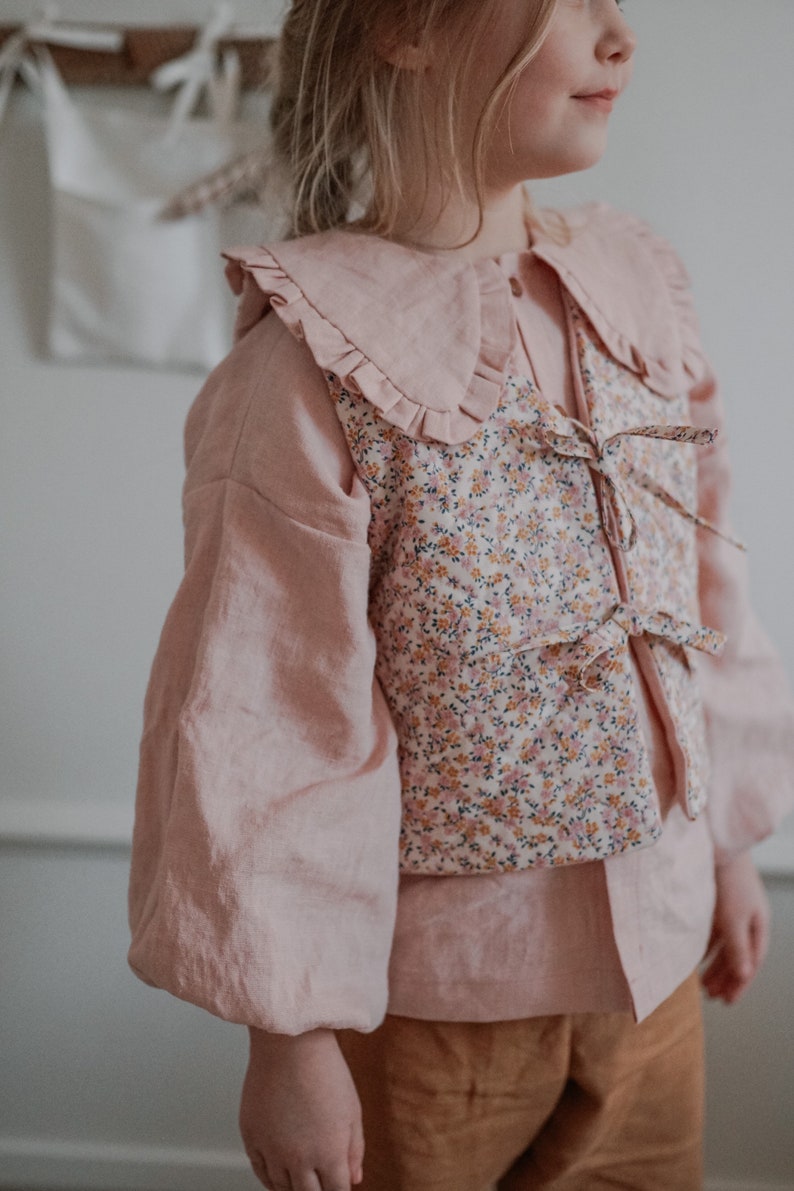 Pu&ograve; includere: Una blusa rosa con un grande colletto a balze e un gilet a stampa floreale. Il gilet ha un nodo sul davanti.