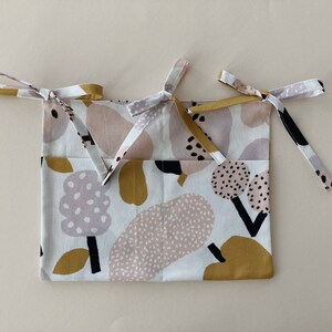 May include: A white fabric storage bag with a colorful abstract print in shades of pink, brown, and black. The bag has two long ties that can be used to hang it.
