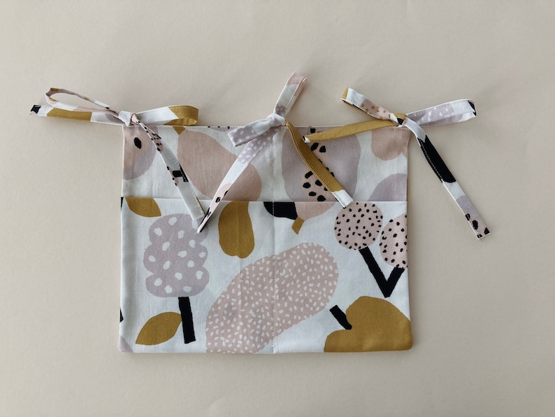 May include: A white fabric storage bag with a colorful abstract print in shades of pink, brown, and black. The bag has two long ties that can be used to hang it.