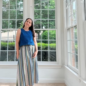 May include: A sleeveless denim blue top paired with a long skirt featuring vertical stripes in various colors. The skirt has a drawstring waist. The person is wearing woven flats. The outfit is in a bright room with large windows.