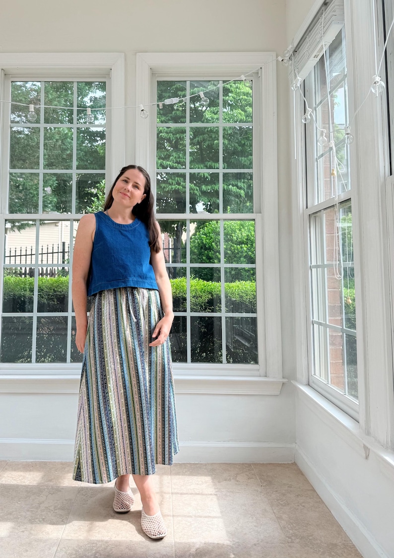 May include: A sleeveless denim blue top paired with a long skirt featuring vertical stripes in various colors. The skirt has a drawstring waist. The person is wearing woven flats. The outfit is in a bright room with large windows.