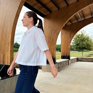 May include: A white eyelet shirt with short sleeves and a collared neckline, worn with dark blue denim trousers. The shirt features a perforated pattern. The person is walking under a wooden structure with a concrete floor.